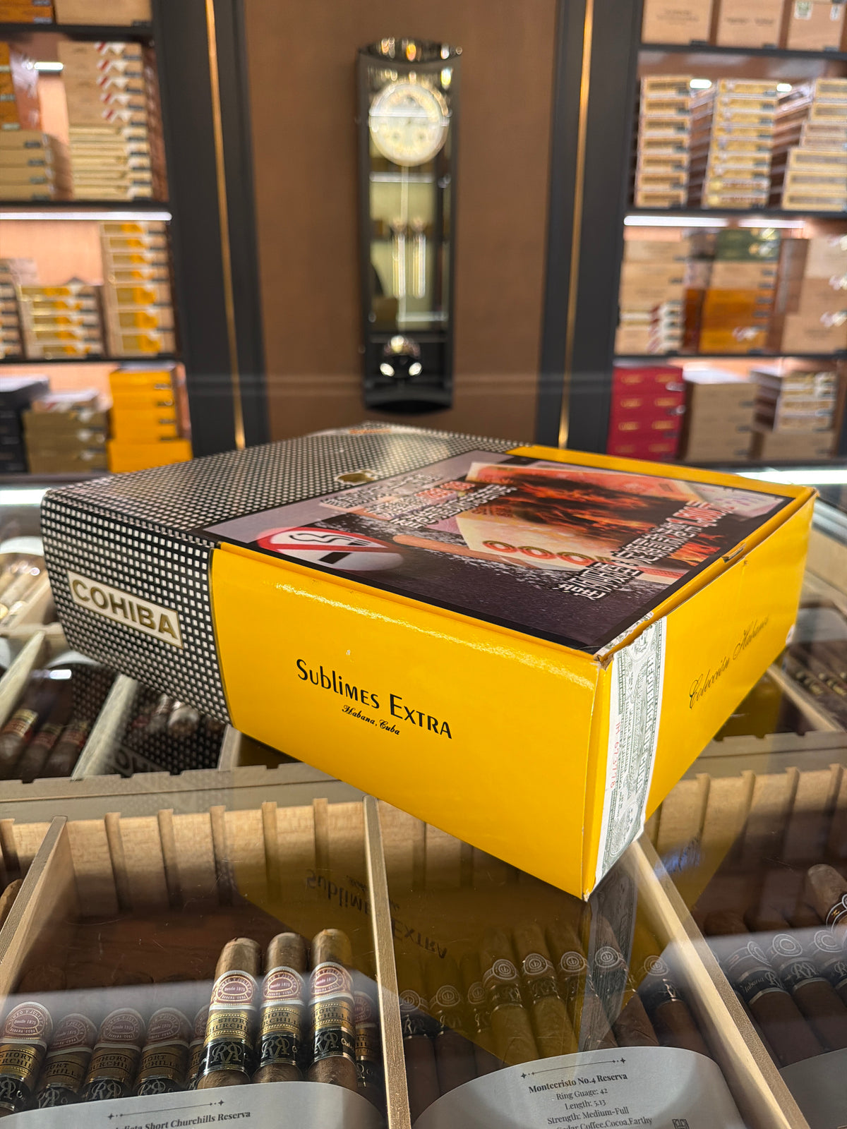 A yellow and black Comba Sublimes Extra cigar box displayed on a glass counter inside a cigar shop, with cigars and shelves visible in the background.