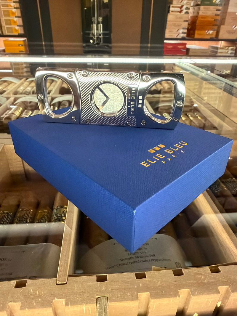 HAMILTON ELIE BLEU cigar cutter in polished stainless steel with diagonal line engraving, resting on a blue gift box.