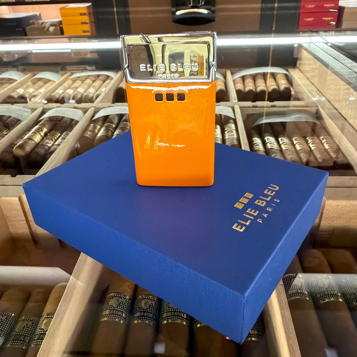 An orange and silver Elie Bleu Paris cigar lighter stands on a blue branded box inside a cigar display case.