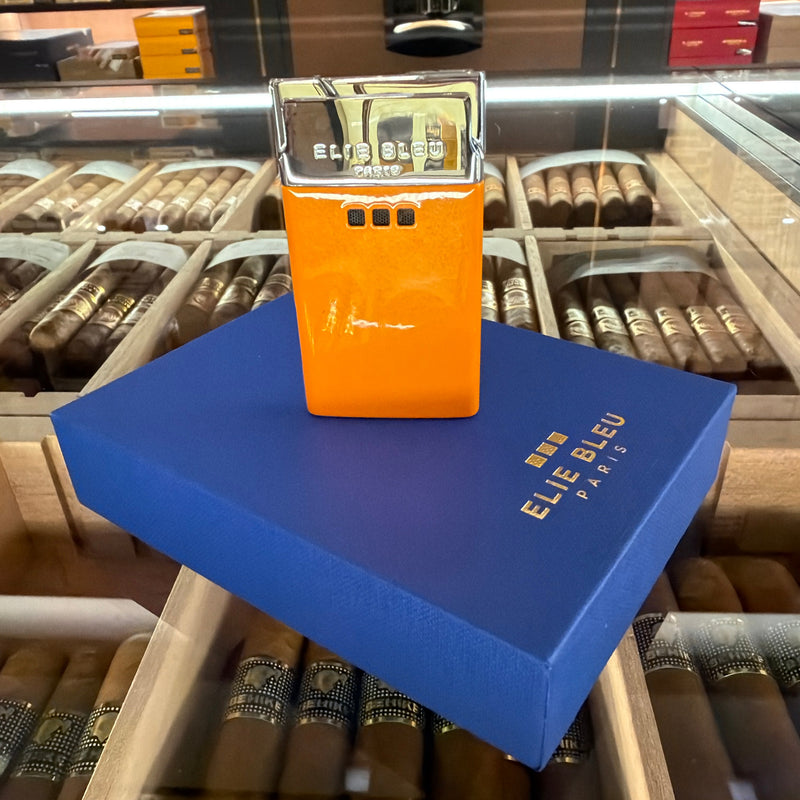 An orange and silver Elie Bleu Paris cigar lighter stands on a blue branded box inside a cigar display case.